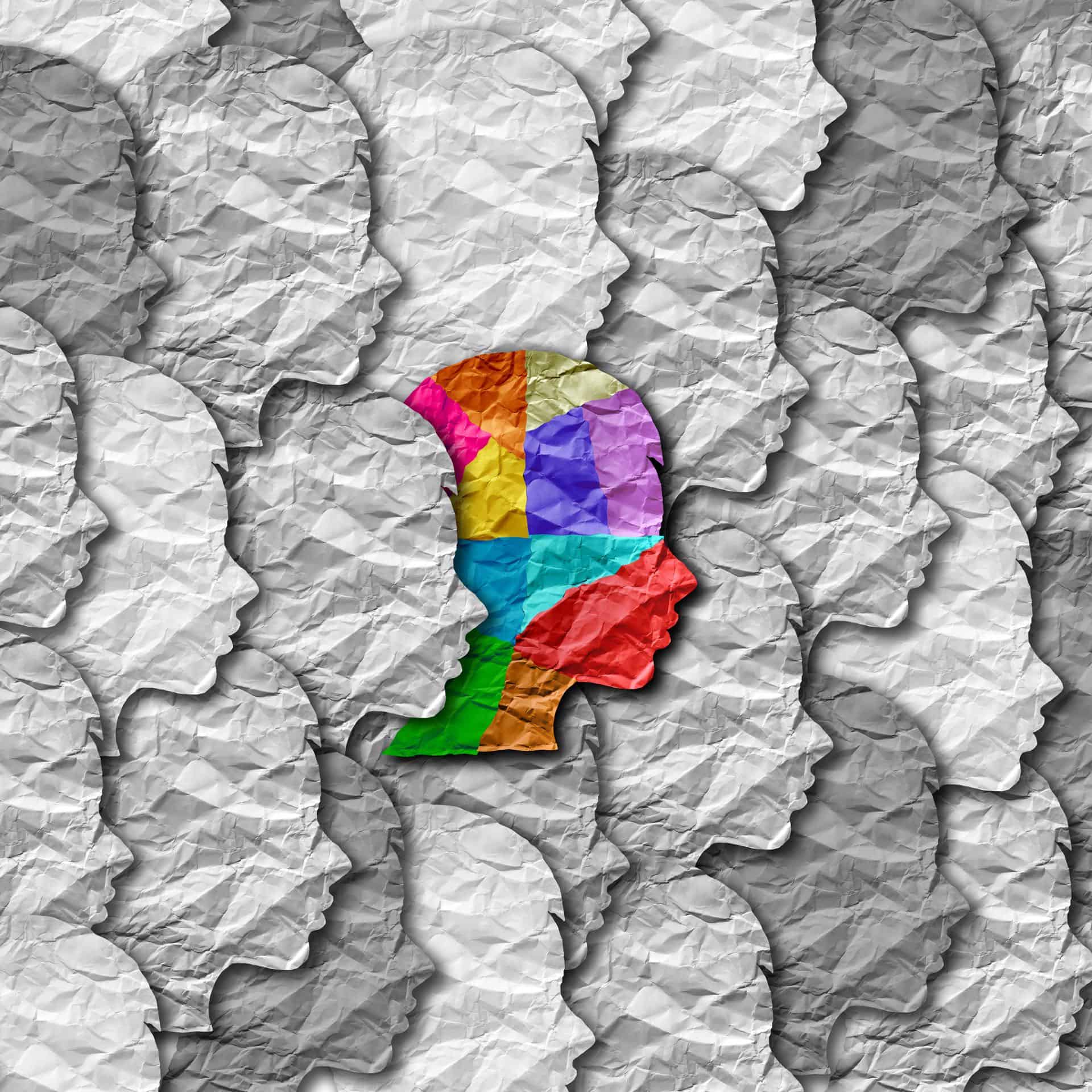 Een kleurrijk silhouet van een menselijk hoofd, samengesteld uit verschillende gekleurde vlekken, valt op tegen een achtergrond van meerdere verfrommelde witte papieren silhouetten van menselijke hoofden naar links gericht. Het beeld symboliseert individualiteit en diversiteit te midden van uniformiteit.
