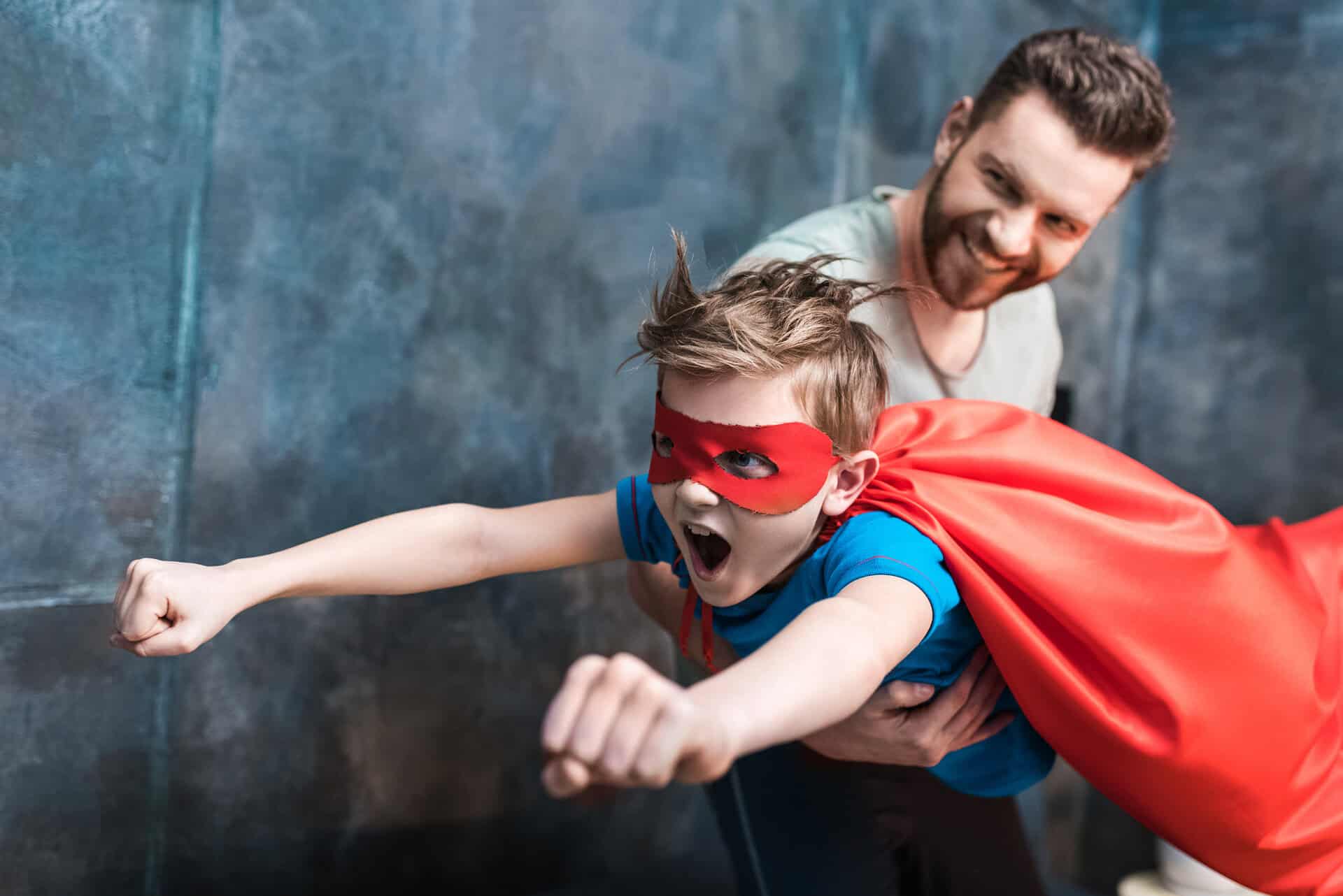 Een jonge jongen, die ADHD lijkt te hebben, draagt een rode cape en een masker terwijl hij wordt opgehouden door een volwassen man, die een vlucht simuleert. De jongen heeft zijn armen uitgestrekt in een superheldenhouding met een opgewonden uitdrukking, terwijl de man lacht en hem van achteren ondersteunt tegen een blauwgrijze achtergrond.